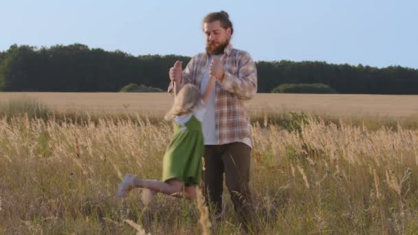 Joven papá caucásico hombre padre papá sosteniendo bebé hija niña de brazos gira giros niño girando en el aire juega juego en el campo de trigo con los niños al aire libre, concepto de unión familiar — Vídeos de Stock