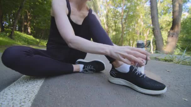 Gros plan jambes féminines de coureuse de jogging mince femme en tenue de sport noire assis sur la piste dans le parc près de la bouteille d'eau après le jogging du matin redresse les liens lacets sur les baskets se préparer pour le jogging de course — Video