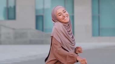 Geleneksel tesettürlü, omuzlarıyla dans eden, kollarını sallayarak mutlu gülümseyen genç Arap kadın portresi. Başarılı İslamcı kız İslami Arap bayan açık havada dans ediyor.