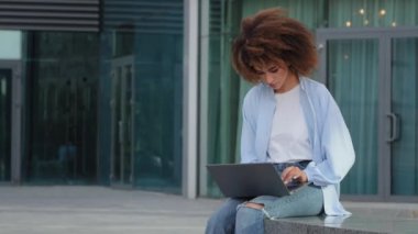 Kıvırcık Afrikalı Amerikalı kız öğrenci serbest çalışan kadın kullanıcı şehir binasının yakınındaki bir sokakta oturuyor. İnternette bilgisayarla uzaktan internet üzerinden yazma kullanıyor. Kablosuz bilgisayar kullanarak.