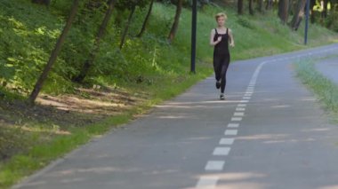 İnce, aktif, orta yaşlı, atletik, beyaz kadın koşucu bayan koşucu. Parkta siyah spor giyer. Yaz sabahı açık havada koşar.