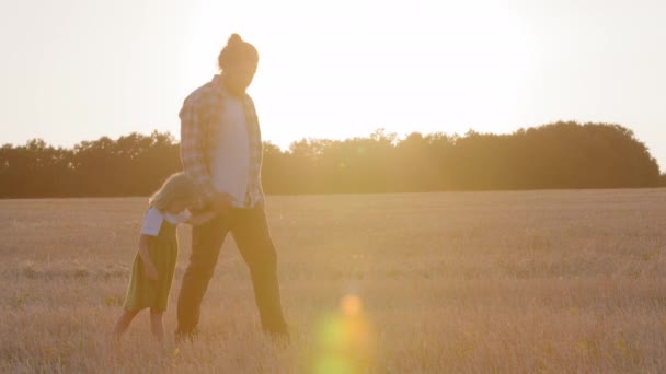 Two silhouettes family parent father dad man farmer holds hand of child little daughter girl small kid walk together across wheat field against background of sunset sunbeams talking walking going — Stock Video