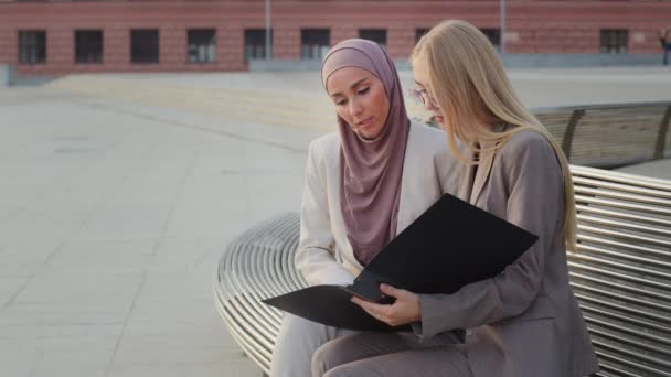 Allvarliga affärskvinnor i kostym håller mapp med dokument, diskuterar rapport. Olika kvinnliga studenter Indiska hijab flicka och ung europeisk kvinna bär glasögon rådgivning under pausen — Stockvideo