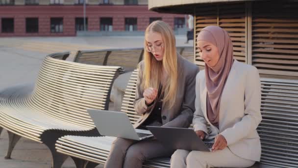 Twee verschillende zelfverzekerde glimlachende collega 's, Indiaas meisje in hijab en jonge Scandinavische Europese vrouw in formele kleding zittend met laptops buiten. Ondernemers delen ideeën of businessplan — Stockvideo