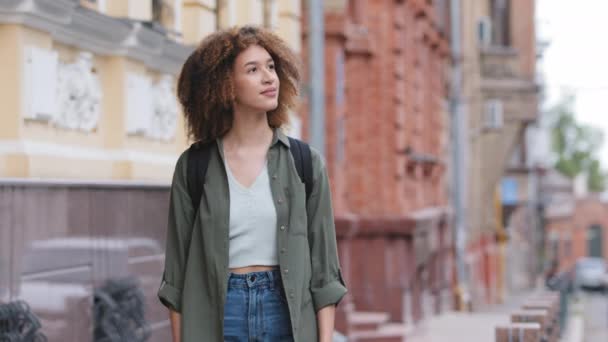 Jovem estudante milenar de raça mista com cabelo encaracolado vestindo roupas casuais andando no centro da cidade. Mulher afro-americana atraente viajando sozinha na cidade admira emocionalmente a arquitetura moderna — Vídeo de Stock