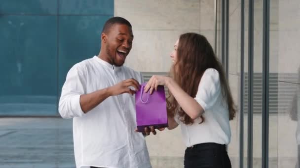 Couple multiracial debout près du bâtiment de la boutique en plein air femme caucasienne montrant l'achat de sac-cadeau à l'homme afro-américain gars regarde à l'intérieur se réjouit bonheur surprise emporter paquet argumenter rire — Video