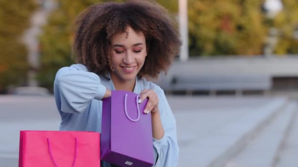 Цікаво, що афроамериканка, афроамериканка кучерява жінка-покупець, яка сидить на вулиці і дивиться на сумочки з подарунками, виглядає у пакунках радісними подарунками. — стокове відео
