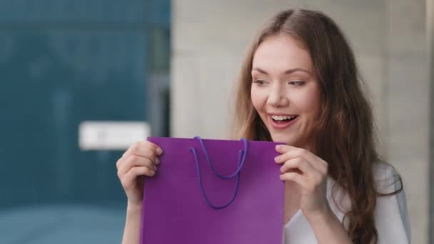 Retrato surpreso surpreso animado caucasiano milenar mulher aniversariante de pé ao ar livre fundo do edifício da cidade mostra segurando saco de presente roxo olha para dentro presentes alegrias sentindo felicidade — Vídeo de Stock