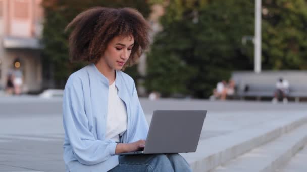 Afro amerikansk ung pige bruger sort blandet race etnisk business kvinde kvindelige studerende manager freelance arbejdstager sidder på fortovet udendørs i byen arbejder med laptop e-learning online browsing hjemmeside – Stock-video