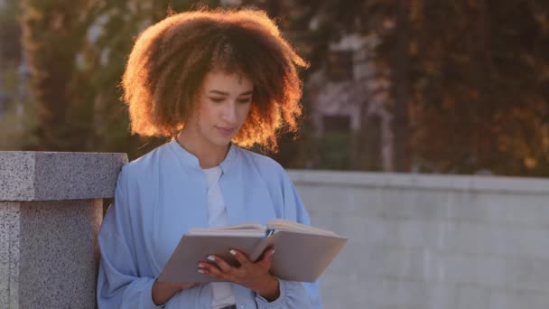 Afro amerikansk kvinna ung afrikansk flicka student läser bok sitter på gatan utomhus i solljus solnedgång njuter litteratur historia vänder sida läsa lära sig information tittar in i anteckningsbok — Stockvideo