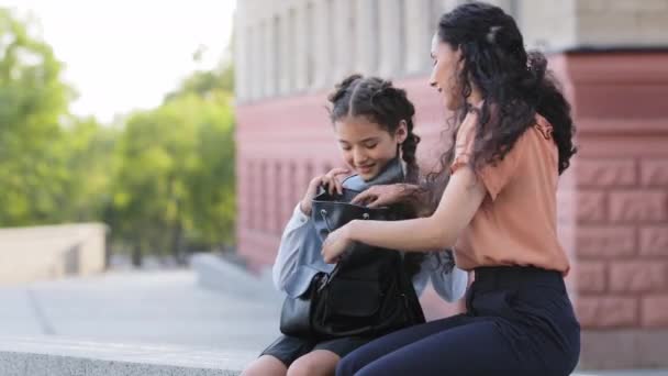 La madre hispana con su hija bebé se sienta al aire libre preparándose para la clase escolar. Mamá ayuda a colegiala niña niña niño estudiante alumno con la preparación para la lección pone cuaderno en charlas bolsa de escuela — Vídeos de Stock