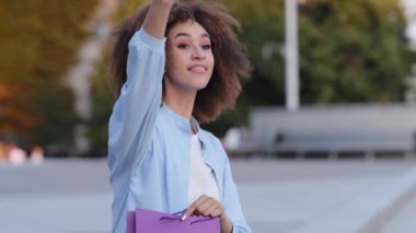 Aktif Afro-Amerikalı Afro-Amerikalı genç bir kadın şehrin dışında oturmuş el sallıyor ve hoş geldin jesti için el sallıyor indirimli alışveriş paketleri sunuyor.