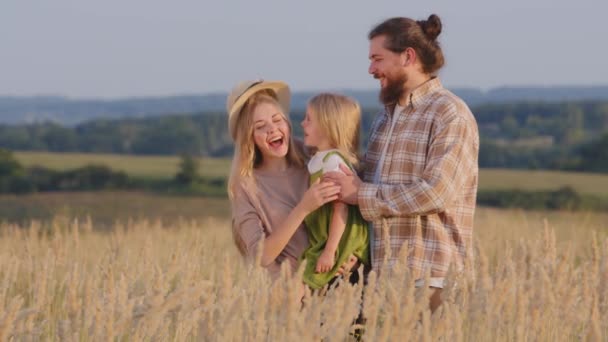 Happy caucasian family stands in wheat field in nature outdoors mother holds little girl daughter father takes off hat from wife puts on baby laughing smiling talking having fun on summer day vacation — Stock Video