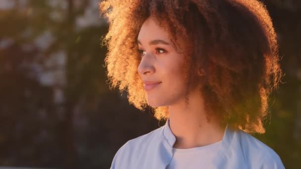 Kadın açık hava portresi güzel etnik Afrikalı Amerikalı kız yan yana gülümseyen kıvırcık saçlı model kadın güneşte gün batımında sokakta kameraya bakarak gülümsüyor. — Stok video