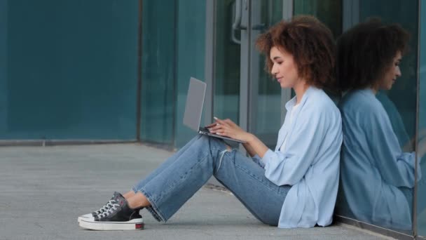 Enfocado joven afroamericana mujer de negocios estudiante chica mujer freelancer usuario sentado en el suelo en la calle al aire libre cerca de edificio de la ciudad de aprendizaje de trabajo con el ordenador portátil de escritura en línea de chat de navegación — Vídeos de Stock