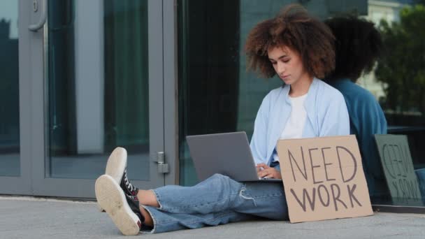 Sérieux afro-américaine femme noire afro fille assise sur le trottoir sur la rue près du bâtiment de verre près de bannière besoin de travail dactylographier sur ordinateur portable à la recherche de postes vacants en ligne, concept de licenciement du chômage — Video