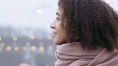 Afro kıvırcık saçlı çekici bir kız, şık modaya uygun eşarpıyla dışarıda dikiliyor, bakışlarını başka yöne çeviriyor, şehirde kış tatilinin tadını çıkarıyor, kamera flörtüne gülümsüyor, güler yüzlü, mutlu bir kadın yüzü takınıyor.