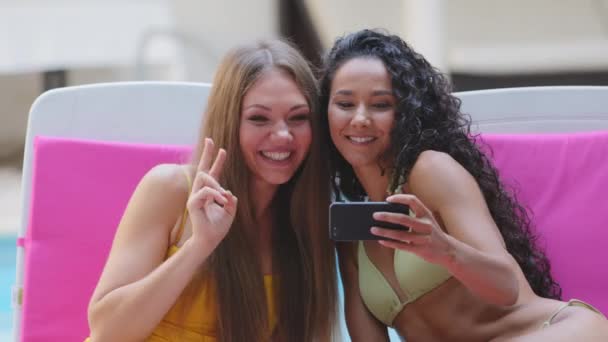 Diverse young women resting on summer vacation sitting on sun loungers near pool having fun. Millennial hispanic European female friends holding smartphone take selfies, show funny facial expression — Stock Video