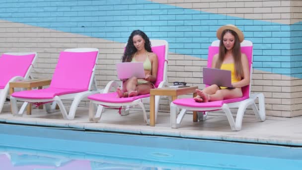 Twee duizendjarige meisjes ontspannen in ligstoelen bij het zomerzwembad. Jonge diverse Latijns-Amerikaanse Europese blanke vrouwelijke vriendinnen zonnebaden buiten met behulp van laptops die op afstand werken. Freelance, afstandsonderwijs — Stockvideo