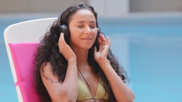 Headshot χαμογελώντας ειρηνική γυναίκα χαλαρώνοντας στην ξαπλώστρα ακούγοντας αγαπημένη μουσική ήχου σε σύγχρονα ακουστικά. Χαρούμενη ευτυχισμένη ισπανόφωνη χιλιετηρίδα κορίτσι απολαμβάνοντας lounge άγχος ελεύθερο χρόνο για τις καλοκαιρινές διακοπές — Αρχείο Βίντεο