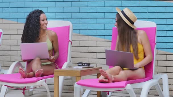 Dwie szczęśliwe, młode, wielorasowe, kaukaskie, latynoskie kobiety, freelancerki siedzące na leżaku, relaksujące się przy basenie i korzystające z laptopów przy basenie podczas wakacji. Praca zdalna w dowolnym miejscu na świecie — Wideo stockowe