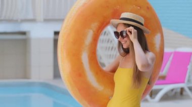 Tasasız, güzel, şık, beyaz tenli, uzun saçlı, şapkalı, güneş gözlüklü, spa otelinde. Mayolu Avrupalı bir kız plaja yürürken elinde turuncu can simidi şişirilebilir yüzük taşır.