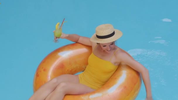 Beautiful girl with exotic beverage in pool. Attractive European blonde young woman model in swimsuit drinking cocktail, floating in orange inflatable swimming lifebuoy, water empty blue background — Stock Video