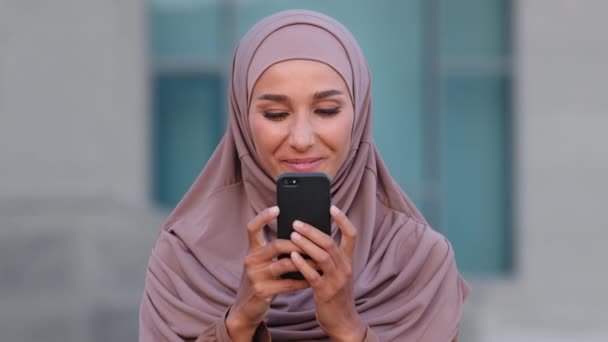 Retrato joven musulmana mujer con estilo hijab pañuelo tradicional de pie al aire libre en la ciudad buscando en la pantalla del teléfono móvil usando smartphone para chatear compras comunicación sonriente en línea — Vídeos de Stock