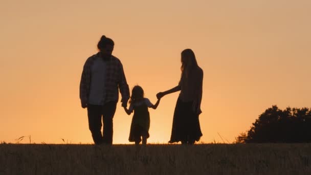 Silueta de grupo de tres personas feliz familia despreocupada padres activos con hija pequeña niña caminando en el campo de trigo saltando caminar en el fondo de los rayos de sol puesta del sol al aire libre en la naturaleza — Vídeos de Stock