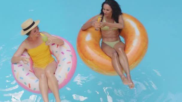 Fröhlich unbekümmert sonnengebräunte Freundinnen mit Cocktails, die den Sommer am Pool genießen. Schlanke Modelle europäisches blondes Mädchen und hispanische junge Frau schwimmen in aufblasbarem Schwimmrettungsring auf dem Wasser