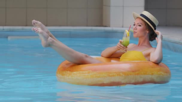 Jolie jolie jeune femme blonde européenne en maillot de bain et chapeau profiter week-end serein avec cocktail dans la piscine seule. Modèle de fille mince millénaire flottant en bouée de sauvetage gonflable orange sur l'eau — Video