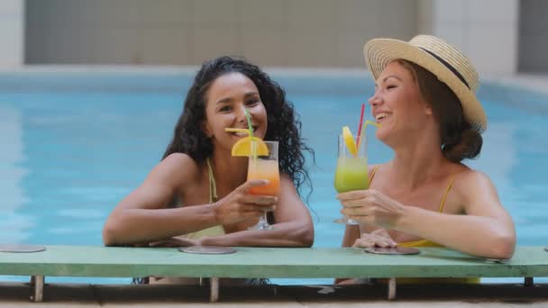 Duas felizes namoradas multirraciais engraçadas segurando coquetéis exóticos de frutas de gelo não alcoólicas, fazendo torcida, copos de clink, beber alegria e celebrar uma reunião amigável perto da piscina de verão no complexo de spa de luxo — Vídeo de Stock