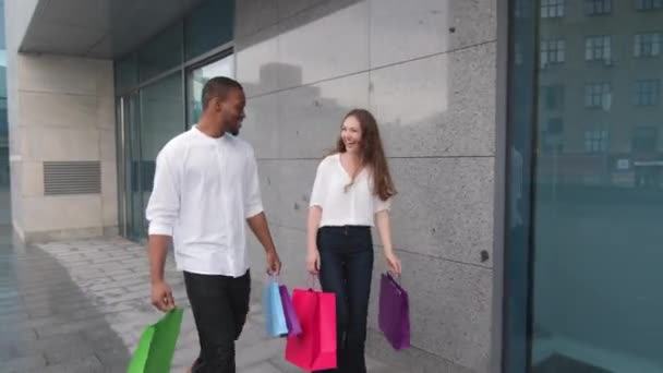 Unga positiva multietniska par lämnar butiken tillsammans med inköp promenader i staden utomhus. Glad diverse afro amerikan man och kaukasiska kvinna gå ut från köpcentret med shoppingväskor talar — Stockvideo