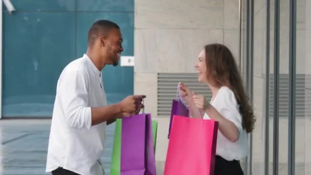 Heureux couple diversifié multiethnique multiracial homme et femme acheteurs shopaholic avoir du plaisir en plein air près du centre commercial danser avec des sacs lumineux achats rire danse joyeuse déménagement en ville — Video