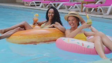 İki çok ırklı, beyaz tenli ve bronzlaşmış, mayo giymiş, mavi suda şişme halkada yüzen iki kadın. Neşeli kız arkadaşlar yaz havuzunda kokteyllerle rahatlıyorlar.