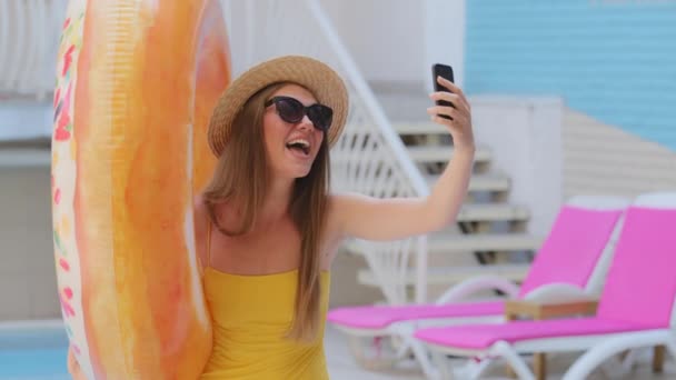 Attractive European blonde girl in swimsuit travel blogger makes selfie, live broadcast, recording video on mobile phone camera near pool. Millennial woman tourist with lifebuoy inflatable ring — Stock Video