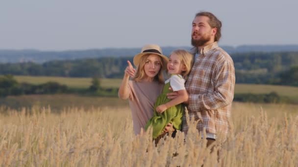 Küçük kızı işaret parmağıyla işaret eden beyaz bir anne buğday tarlasında babasının yanında durduğunu açıklıyor. Aile babası ve kız çocuğu doğada duruyor. — Stok video