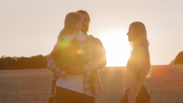 Silhuetas família pais com filha criança stand no campo de trigo fundo de raios de sol pôr do sol pai segura bebê nos braços mãe mostra polegares para cima como aprova andar na natureza ao ar livre, dia de verão — Vídeo de Stock