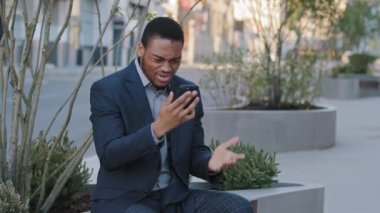 Sinirli Afro-Amerikan siyah iş adamı, elinde akıllı telefonuyla telefon ekranına bakıyor. İş görüşmesi videosu çekiyor. Müzakere sonuçlarından, değişimden, önerilen şartlardan memnun değil.