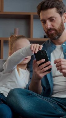 Evde mutlu bir beyaz aile babası ve küçük kızı cep telefonu kullanıp gülümsüyor ve akıllı telefonlu çocuk çocuk babasıyla internetten mal alışverişi yapıyorlar.