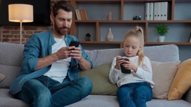 Evde beyaz bir aile, kızı olan bir cihaz bağımlısı babası, cep telefonlu bir çocuk, cep telefonlu bir çocuk, kız çocuğuna akıllı telefon gösteriyor ve birlikte sohbet ediyorlar. Çevrimiçi bağımlılık sosyal medyası.