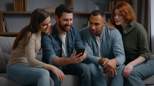 Four multiracial friends at home party looking in phone discuss news app mobile service diversity people using smartphone together on couch multiethnic men women watching funny video talking happy