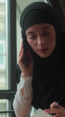 Stresli, çok çalışan, çok görevli, Müslüman iş kadını, tesettürlü, kadın, tesettürlü, iş kadını, konuşan, cep telefonlu, dizüstü bilgisayara bakan, not defterinde veri arayan, iş sıkıntısı çeken.