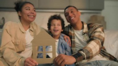 Mutlu gülümseyen Afro-Amerikan ailesi, ellerinde karton ev şekliyle kameraya bakıyor. Kaygısız ebeveynlere bakıyorlar.