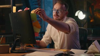 Akşamları sinirli Kafkasyalı iş adamı monitör ofisindeki bilgisayardan çıkartmaları atıyor. Aşırı çalışan iş adamı stresli. Yapışkan notları kontrol ediyor.