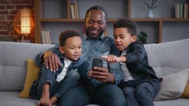Evde akıllı telefonlu mutlu Afrikalı aile kanepesinde küçük oğulları internet karikatürleri gösteren cep telefonu alışverişi yapan babaları e-ticaret babaları çocuklara uygulama kullanmayı öğretiyor