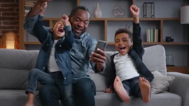 Mutlu Afro-Amerikan ailesi evde telefonla kazanıyor baba iki oğluyla çocuklar mobil kumarhane kazanıyor online zafer kutlanıyor beşlik akıllı telefon sevinci