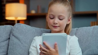 Kafkasyalı gülümseyen mutlu küçük kız, evde ders kitabı yazıyor uzaktan eğitim öğreniyor, ilkokulda ilkokuldan zevk alan kız öğrencileri okuyor, oturma odasında ödevlerini yazıyor.