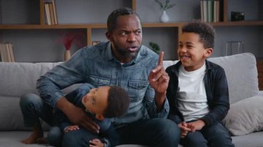 Mutlu Afro-Amerikan ailesi evlerinde kanepede oturup babalarına sarılıyorlar. İki küçük çocukla gülüyorlar. Çocuklar eğleniyorlar.