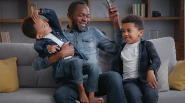 Mutlu Afro-Amerikan ailesi telefonla gülüyor kanepede oynaşıyor evde baba oğul gülüyor çocuklar kucaklaşıyor mobil uygulamalar kullanarak eğleniyorlar neşeli oyunlar oynuyorlar
